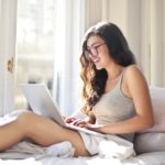 A happy woman in grey sitting on bed using her laptop reading funny profile headlines